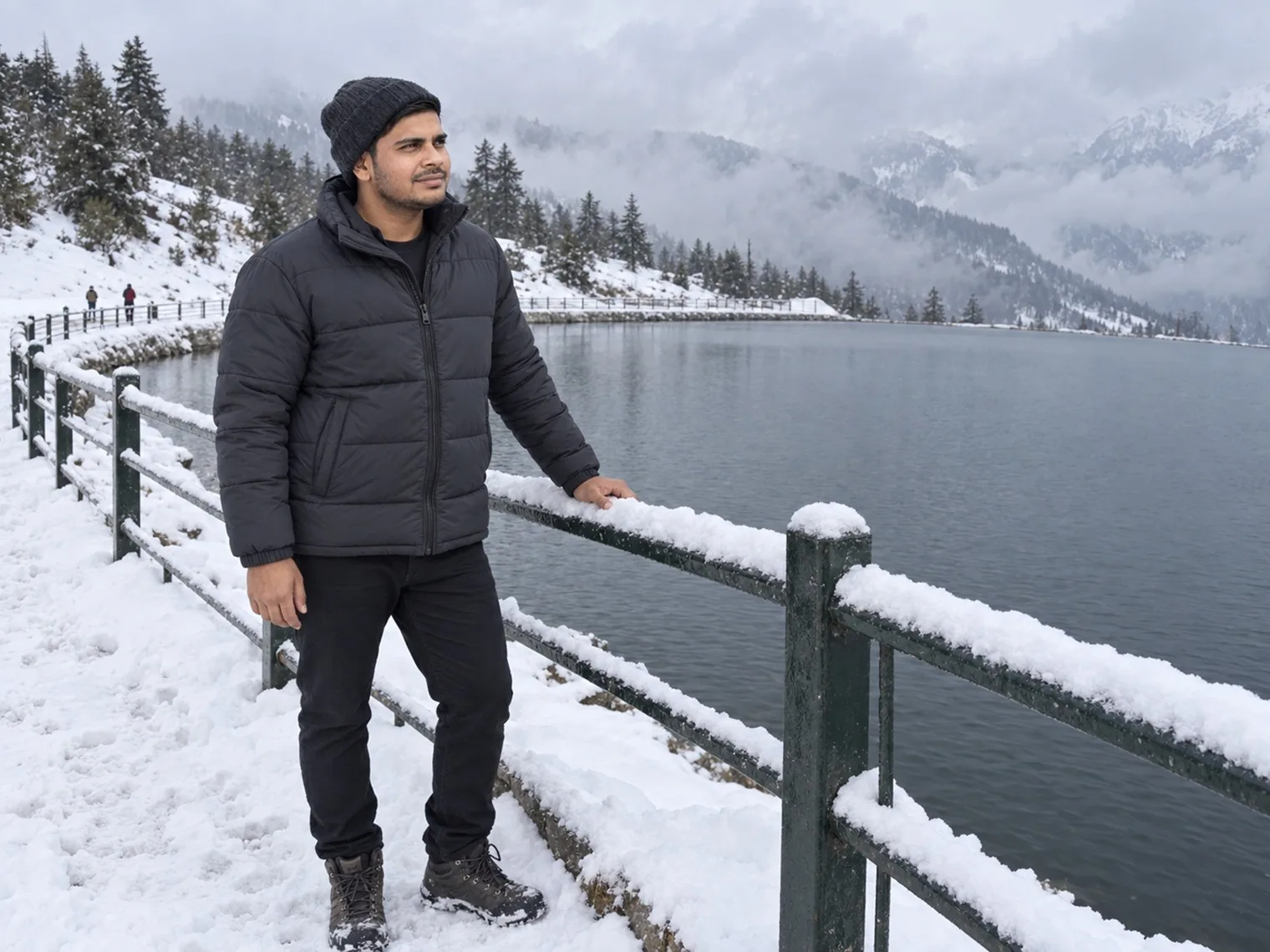 Nav standing by a snow-covered railing beside a large dark reservoir in January 2026, pines and cloud across the water.