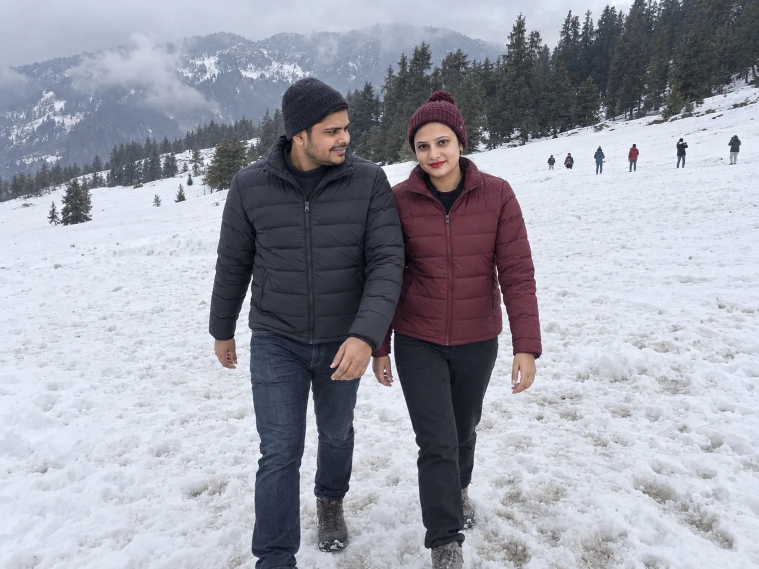 Nav and Vandana walking across deep snow on an open slope, pine trees and cloud behind.