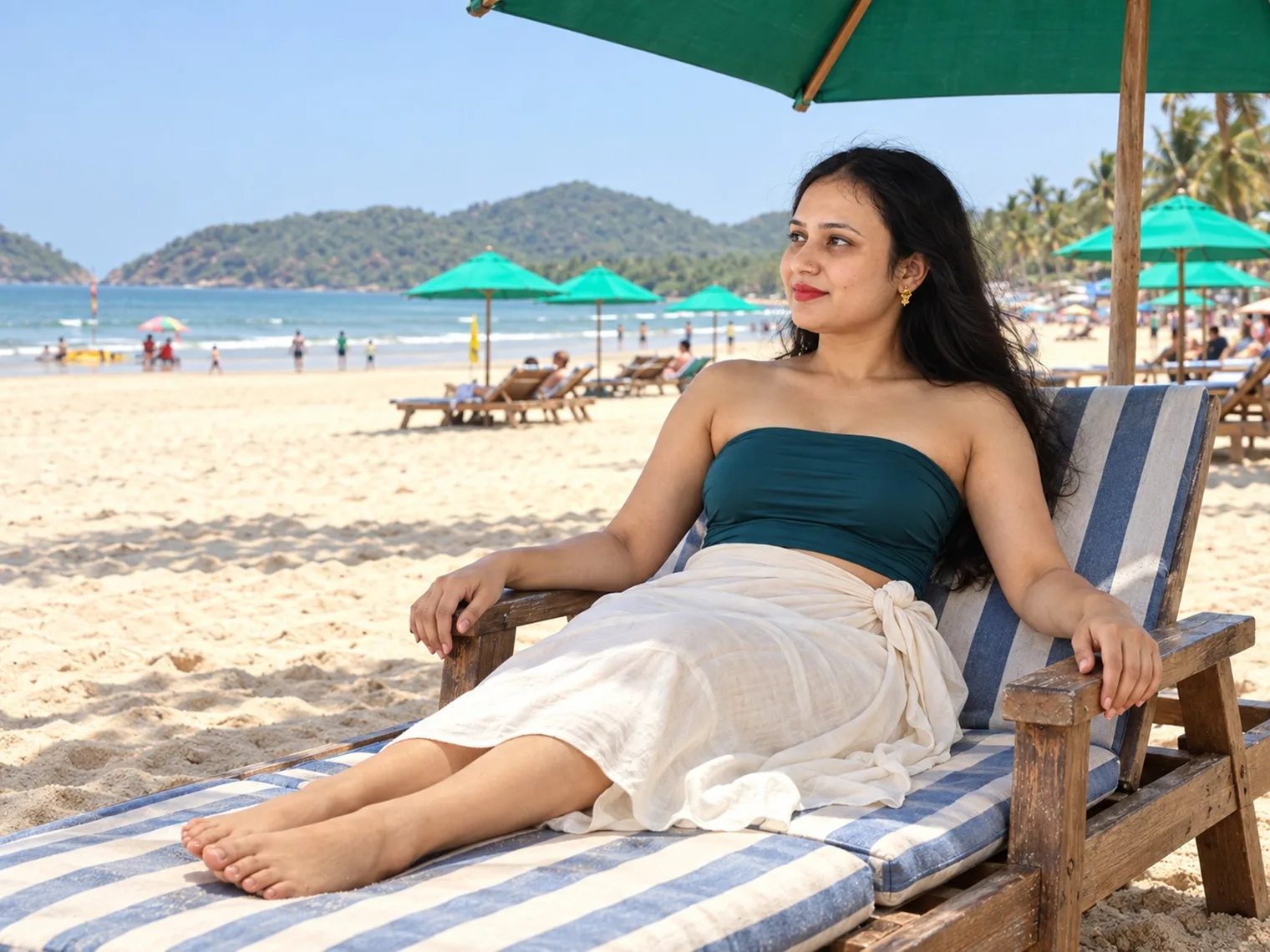 Vandana reclining on a striped lounger under a green umbrella on Palolem beach, more umbrellas, loungers and people along the sand and a wooded headland across the bay.