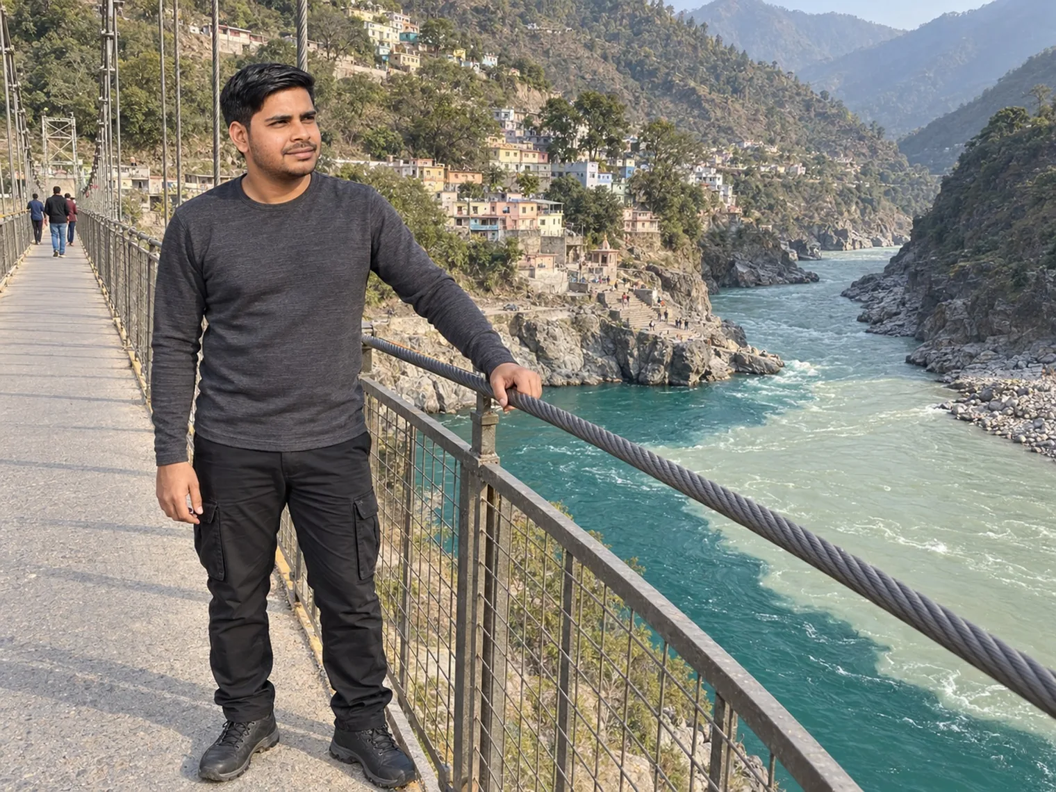 Nav standing on a narrow suspension footbridge above a river, houses stacked on the far bank.