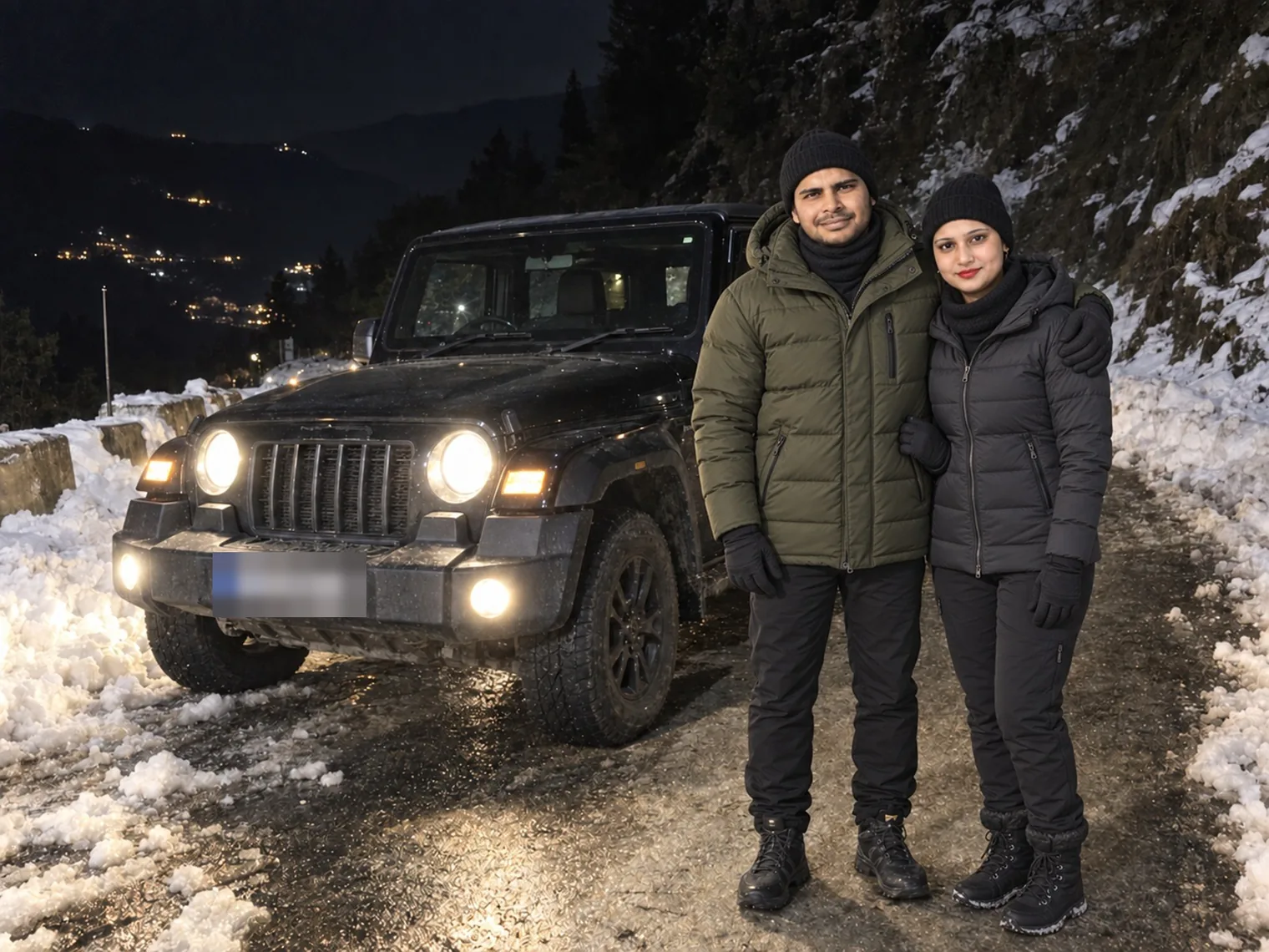 A black Thar with its headlights on beside a snowy bank at night, Nav and Vandana in winter hats standing next to it, town lights far below.