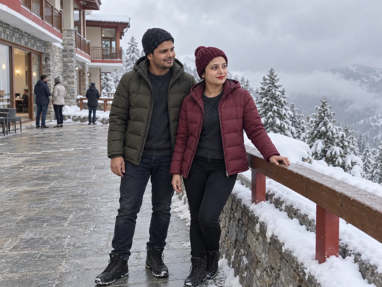 Nav and Vandana beside a snow-covered wooden railing on a stone terrace, hotel balconies to one side and snowy pines and cloud to the other.