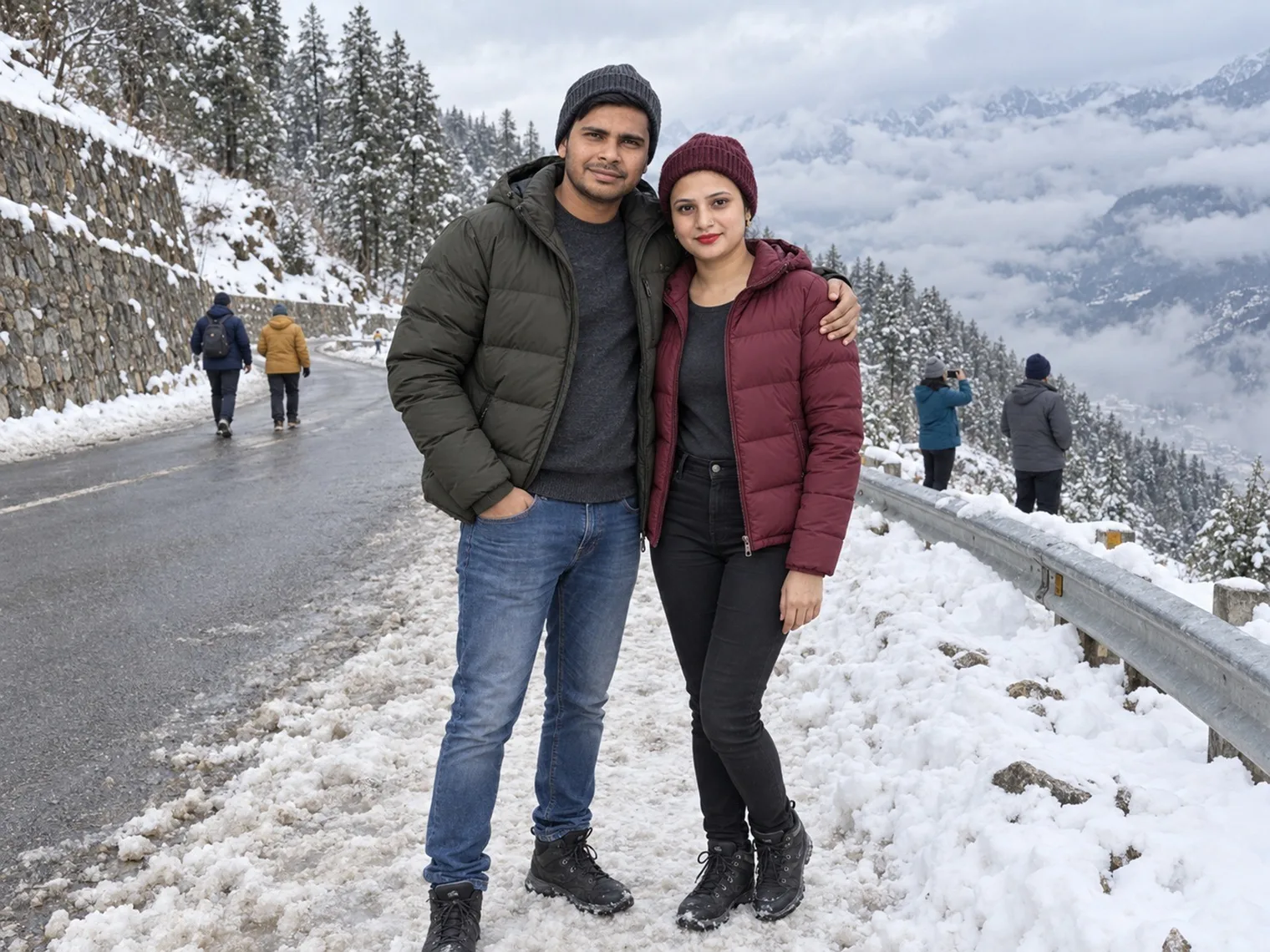 Nav and Vandana in winter jackets and woollen caps standing in packed snow beside a wet mountain road and a metal guardrail, cloud filling the valley below.