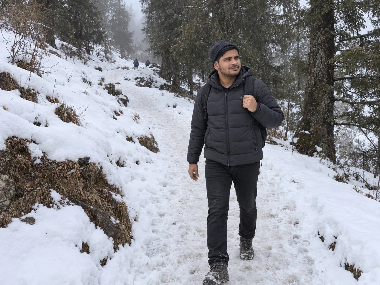 A visitor with a backpack walking along a snowy hillside trail among conifers, a small figure further up the path.
