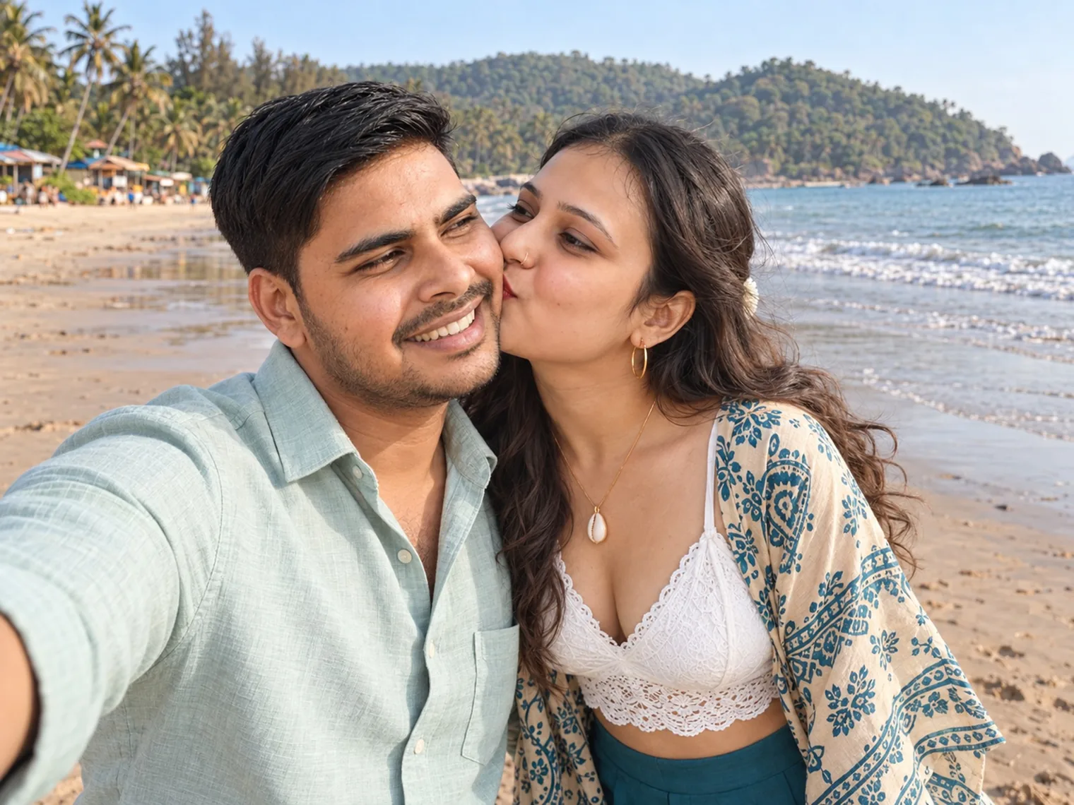 Vandana kissing Nav's cheek in a selfie on Agonda beach, the sea and a green headland behind them.