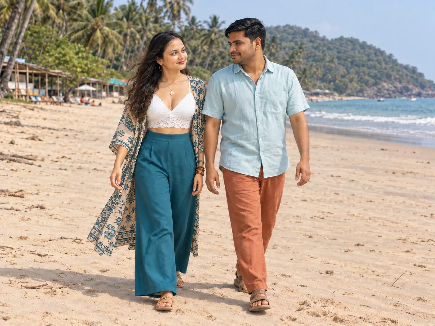 Nav and Vandana walking side by side on the sand at Agonda, palm trees, beach huts and a wooded headland behind them.