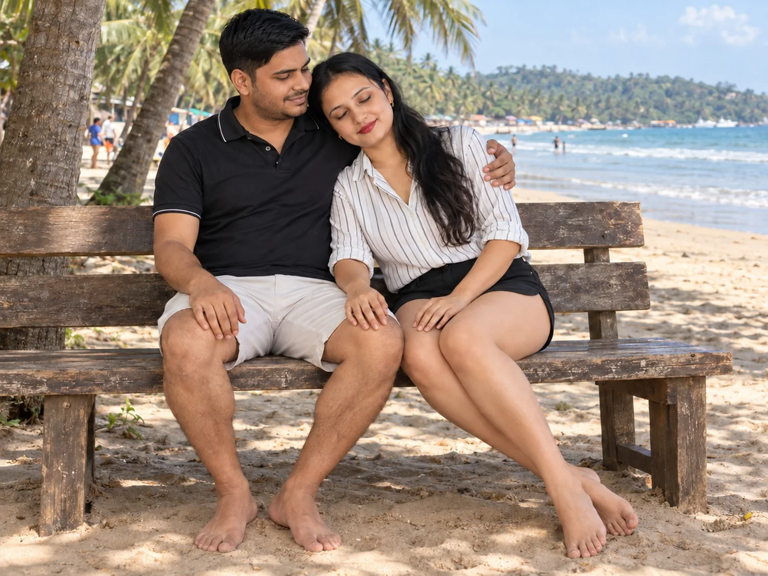 Nav and Vandana sitting close together on a wooden bench in the shade of the palms on Palolem beach, the sea behind them.