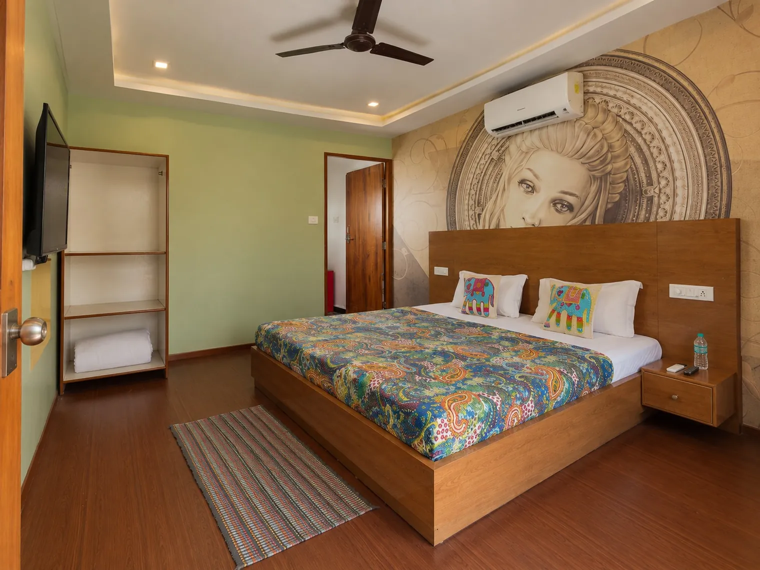 A double room at Palolem Casa: a wooden bed with a patterned bedspread under a large mural of a woman's face, an air conditioner, open shelves and a wooden floor.